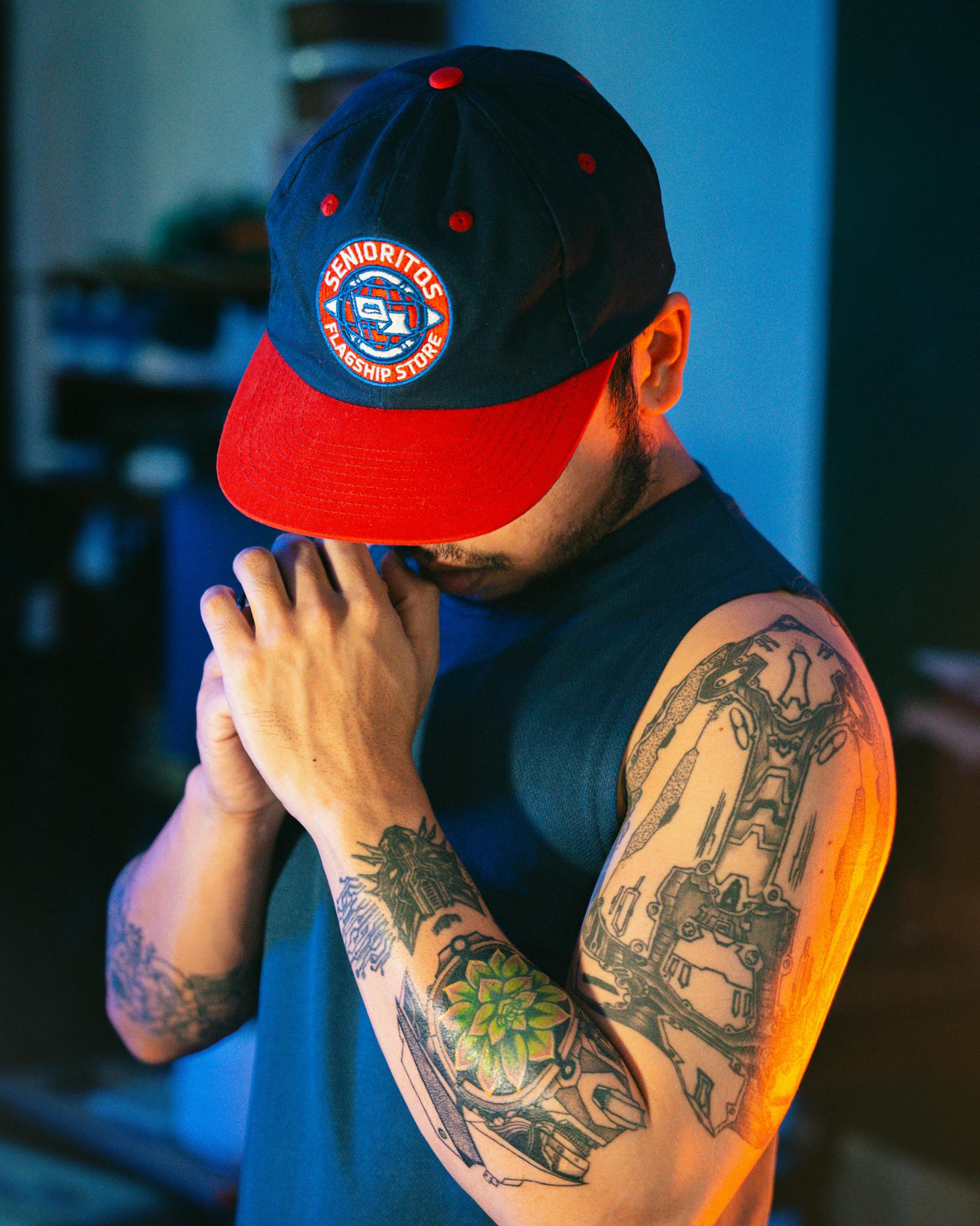 Tattooed man wearing a cap with colorful designs, framed by dramatic lighting.